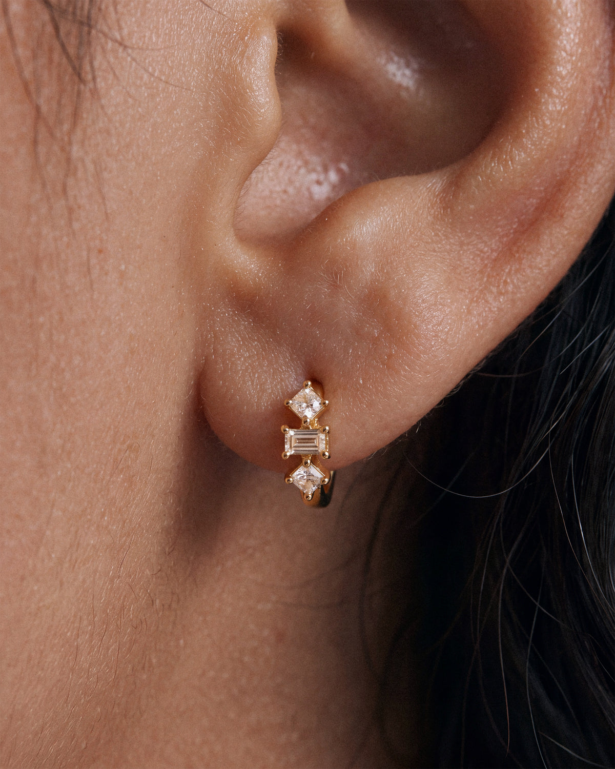 Baguette Trinity Huggie Hoops in 18k Gold worn on the earlobe — super macro close-up of baguette and princess-cut crystal huggie earrings.