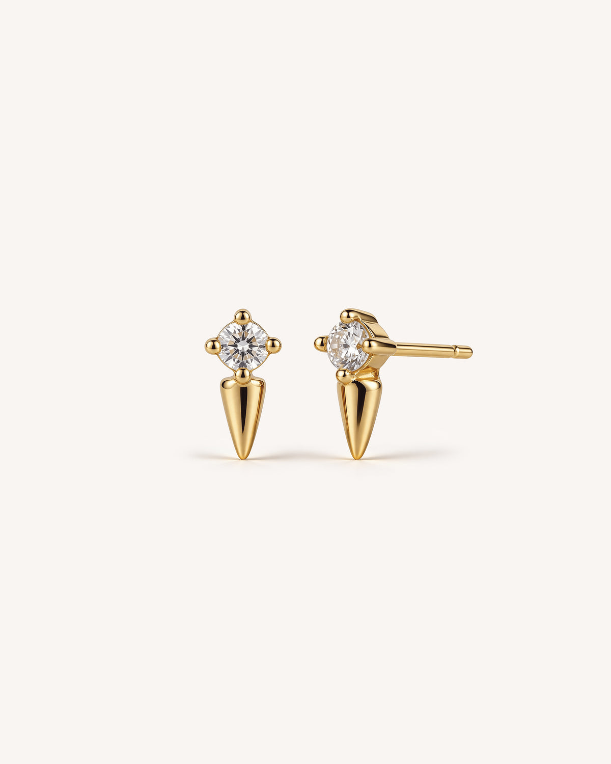 A pair of 18k gold stud earrings with a round-cut clear gemstone in a four-prong setting, mounted on a tapered cone-shaped post, shown against a solid off-white background.