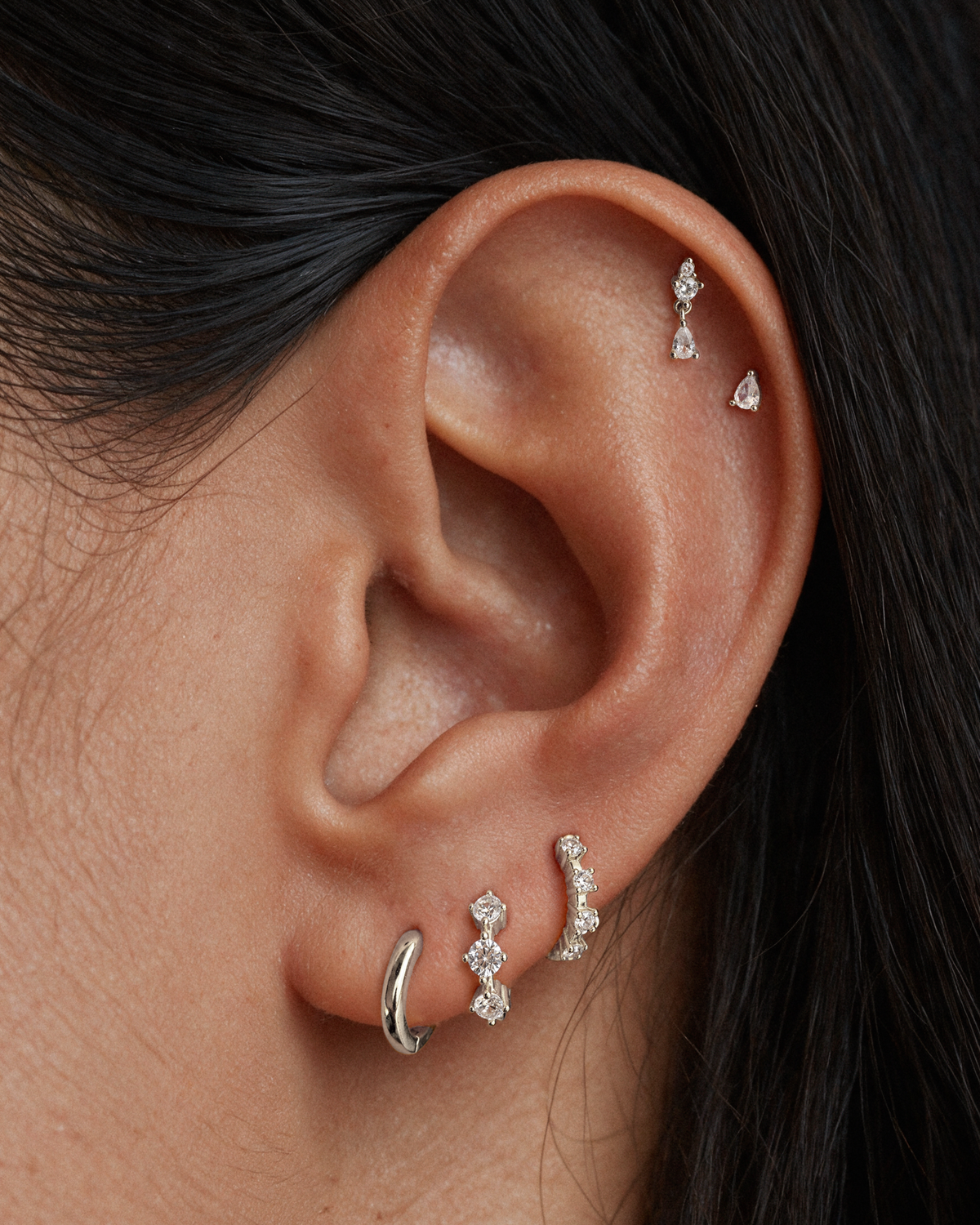 Close-up of a person's ear showcasing multiple 925 sterling silver ear piercings, including a hoop, a curved huggie with clear stones, a marquise stud and one small dangling piercing stud in the upper helix.