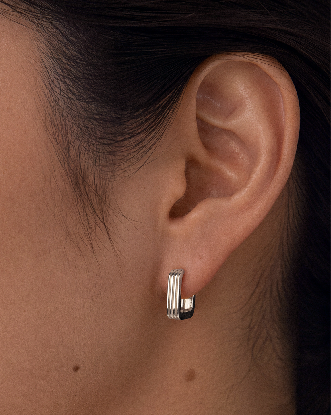 A close-up side profile of a woman wearing a small 925 sterling silver huggie hoop earring with vertical ridges.