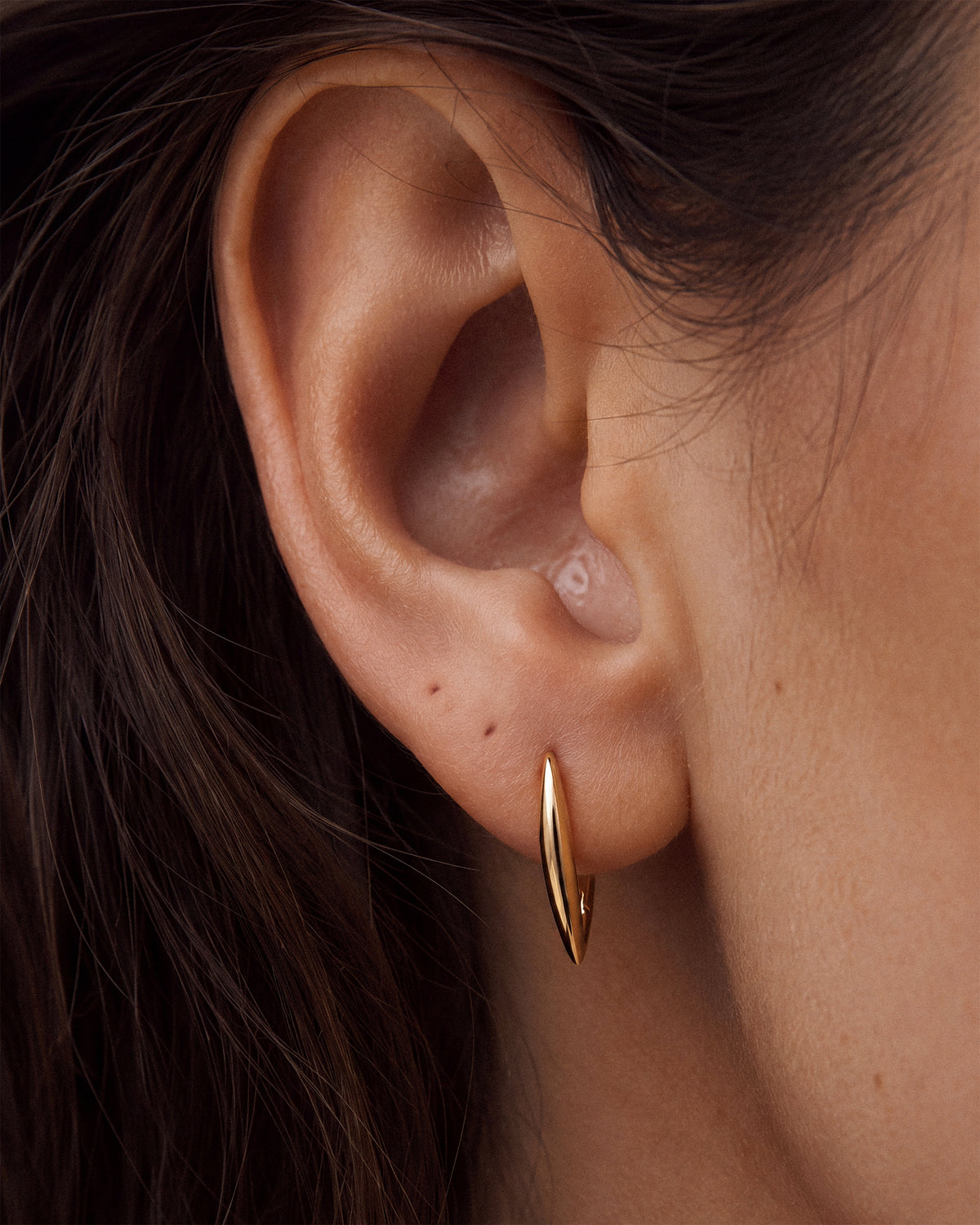 A close-up of a person's ear wearing a thin, tapered 18k gold plated thick micron hoop earring.