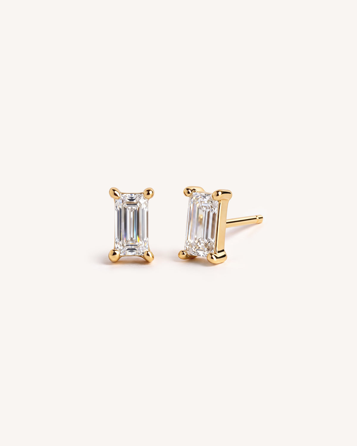 A pair of 18k gold stud earrings with baguette-cut clear gemstones, shown on a clean off-white background.