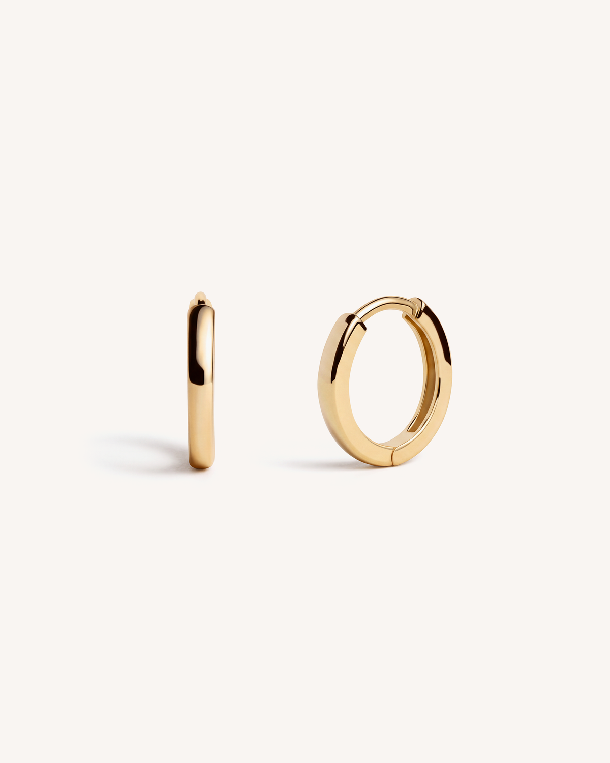 A pair of polished 18k gold plated huggie hoop earrings shown on a clean, off-white background, with one earring standing vertically and the other lying horizontally to display its shape and reflective finish. One of our bestsellers. An essential for every stack.