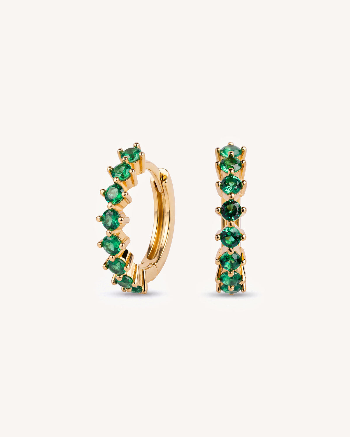 A pair of tennis gold huggie hoop earrings set with round-cut green emeralds, shown on a clean off-white background.