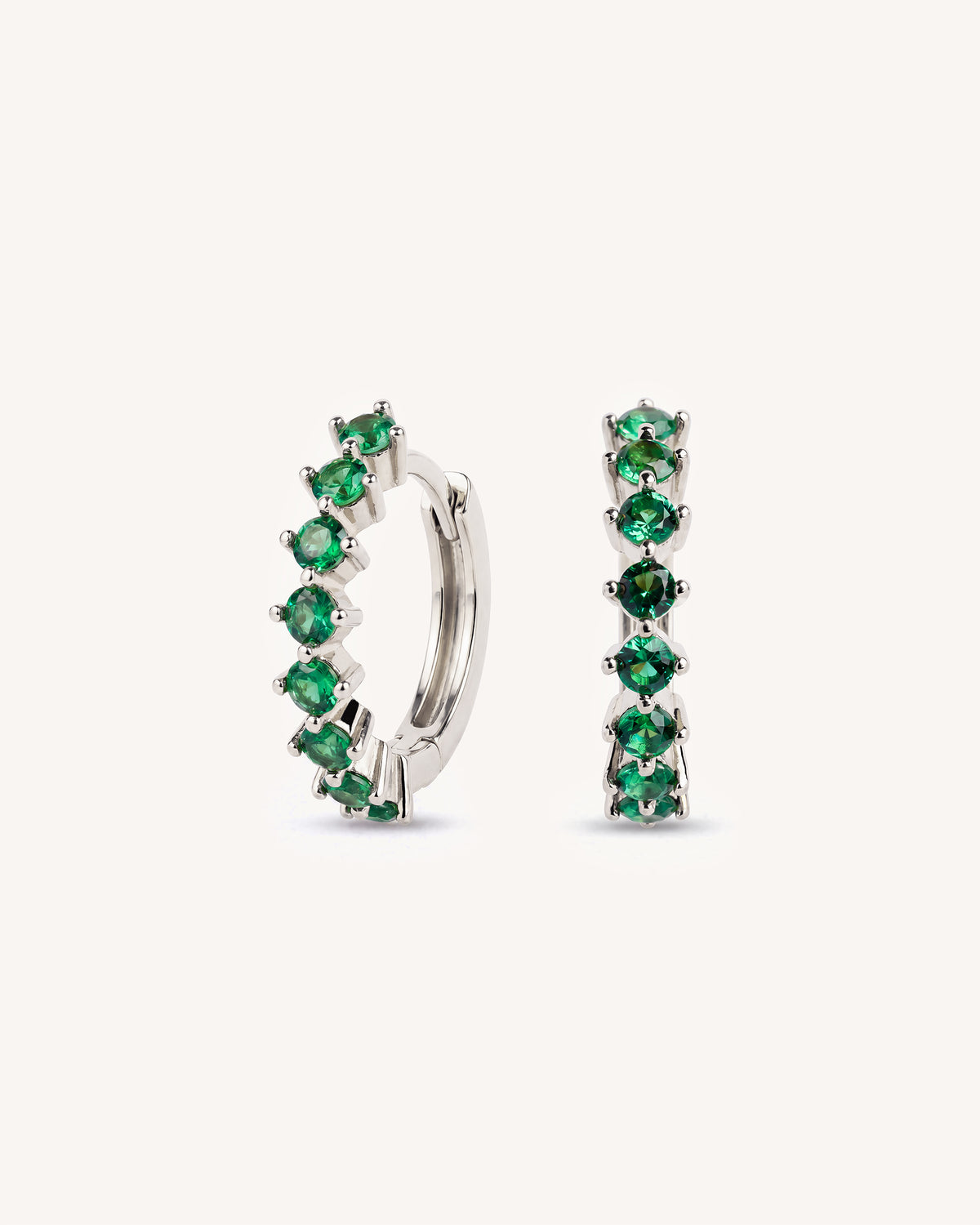 A pair of tennis silver huggie hoop earrings embellished with a row of round-cut green emerald-cut gemstones, shown against a plain off-white background.