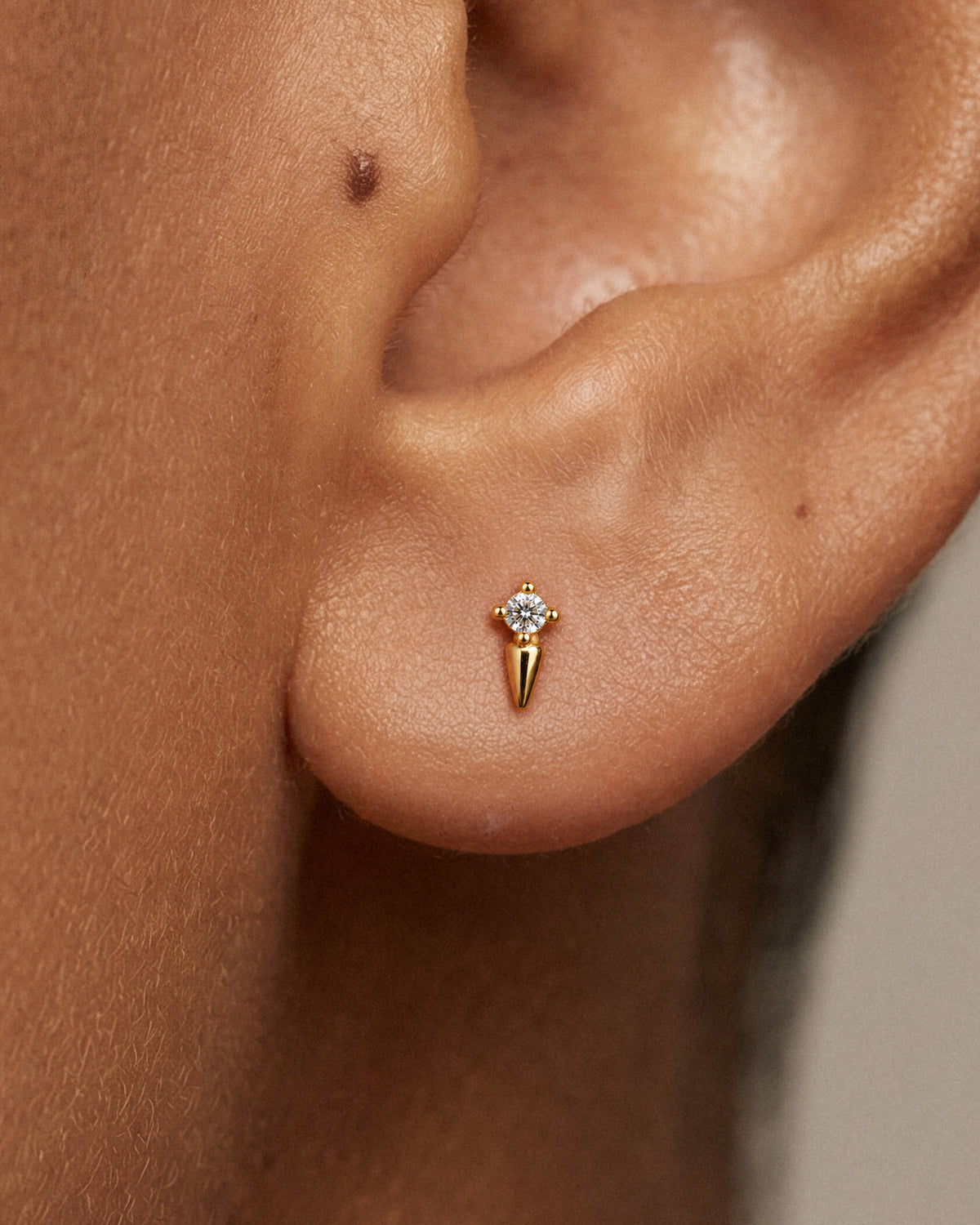A close-up of a model's ear wearing a small 18k gold stud earring with a clear round gemstone and a pointed spike drop below it.