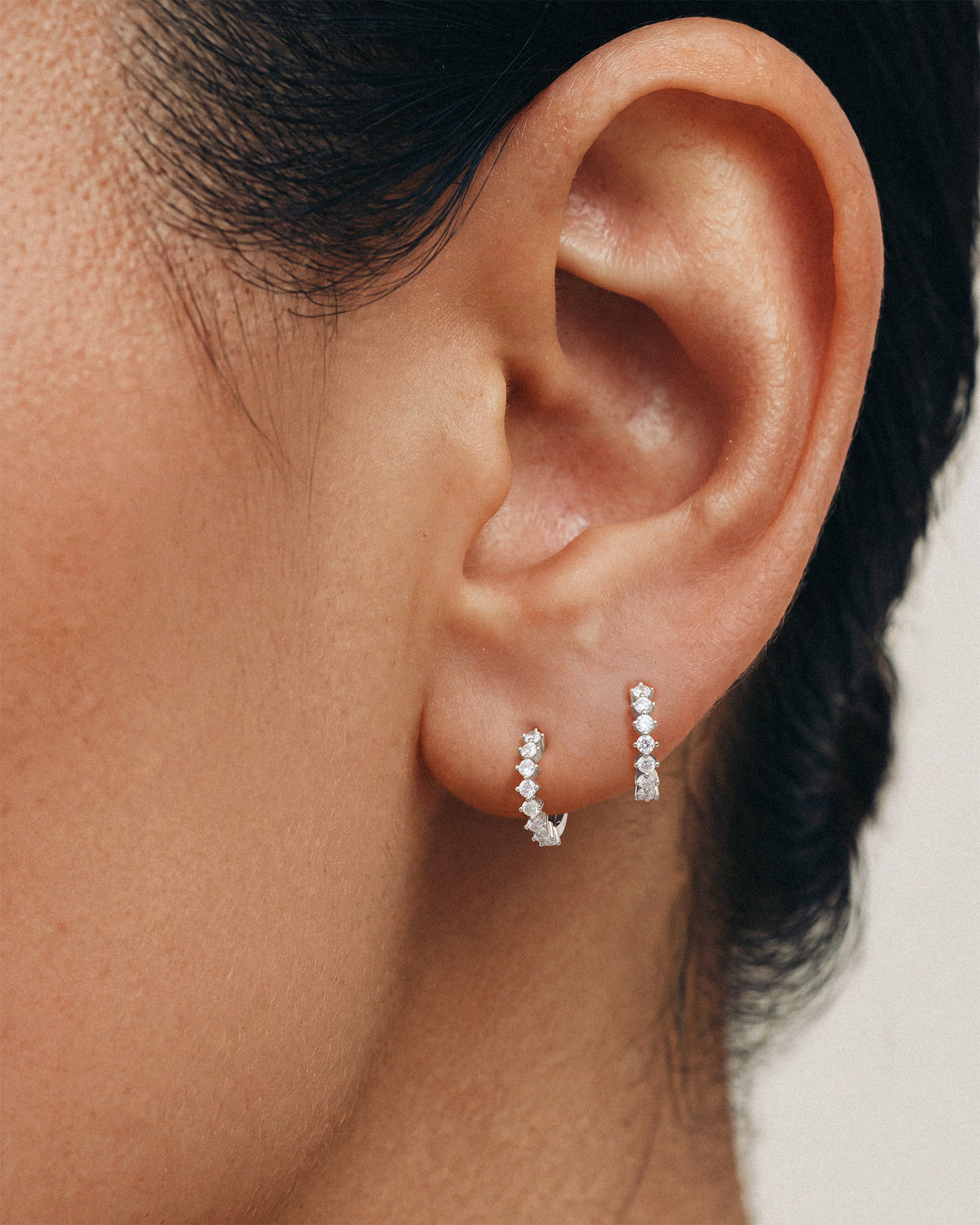 Close-up of a person's ear wearing two small 925 silver hoop earrings with clear stones , against a soft light background.