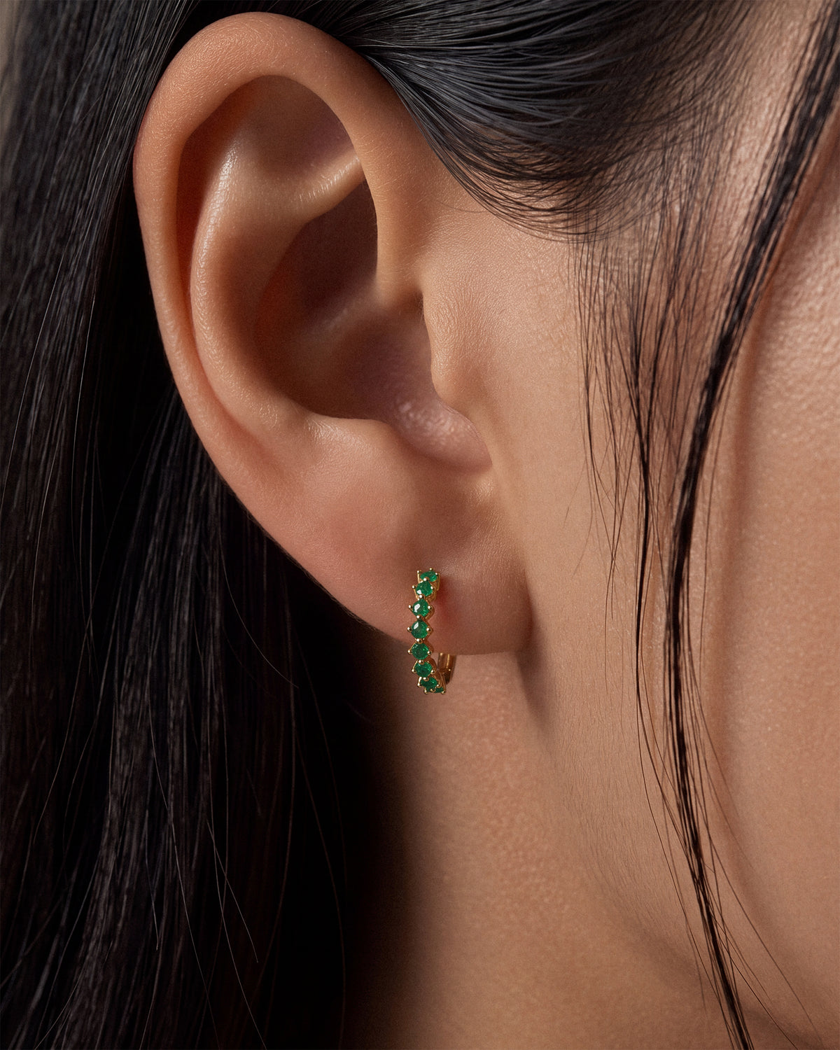 A close-up of a model's ear wearing a small gold huggie hoop earring set with multiple emerald green gemstones.