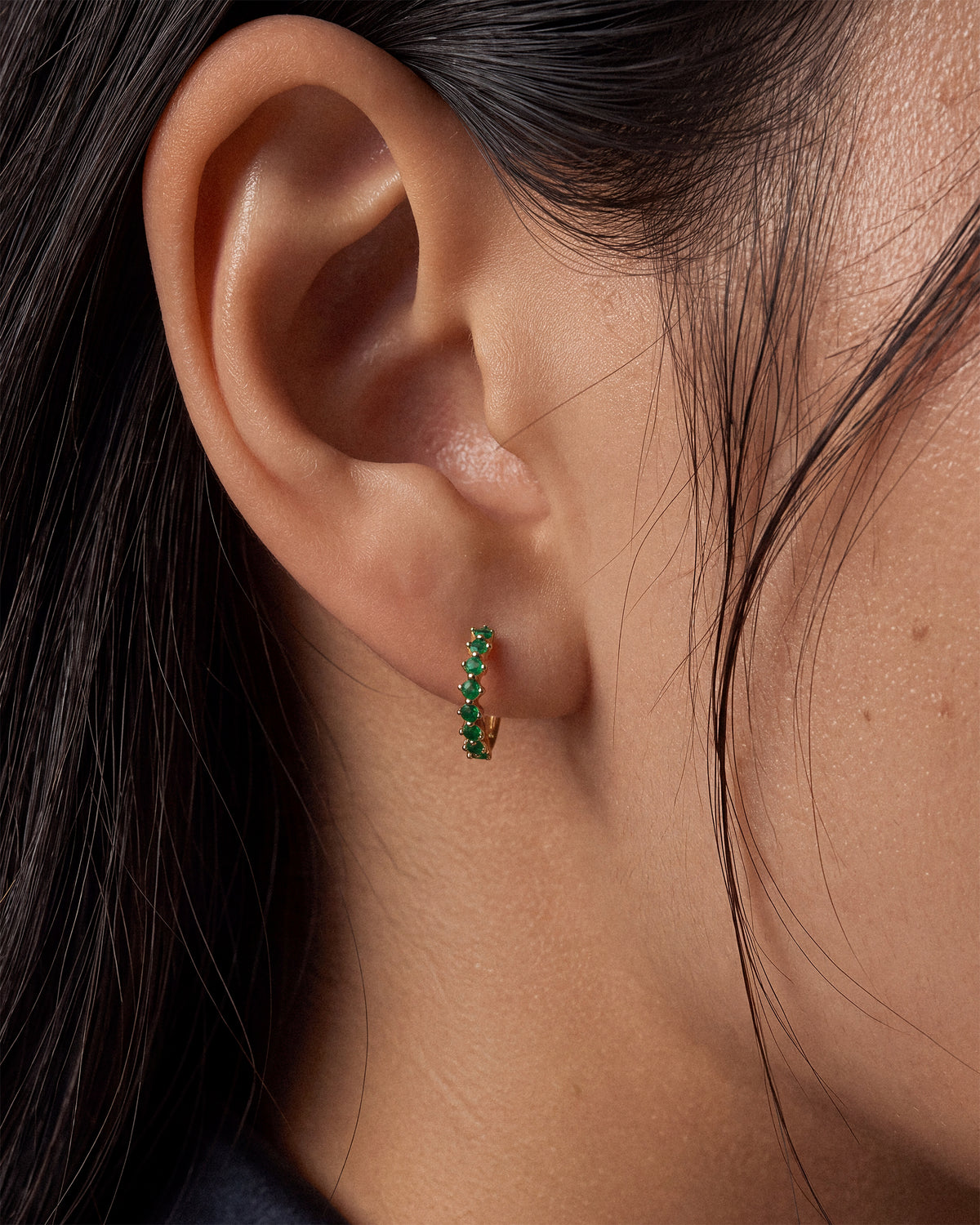 A close-up of a person's ear wearing a small 18k gold plated vermeil hoop earring set with emerald green gemstones.