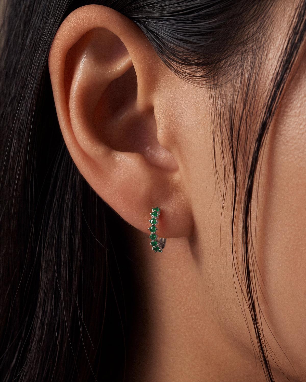 A close-up of a model's ear wearing a small curved 925 silver hoop earring set with emerald green gemstones.