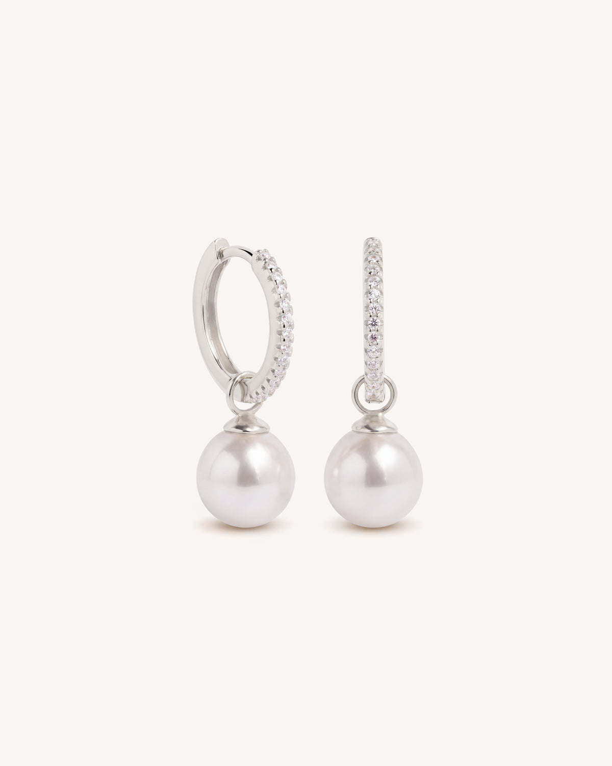 A pair of silver huggie hoop earrings with a row of small clear stones on one side and a dangling white pearl on the other, set against a solid off-white background.