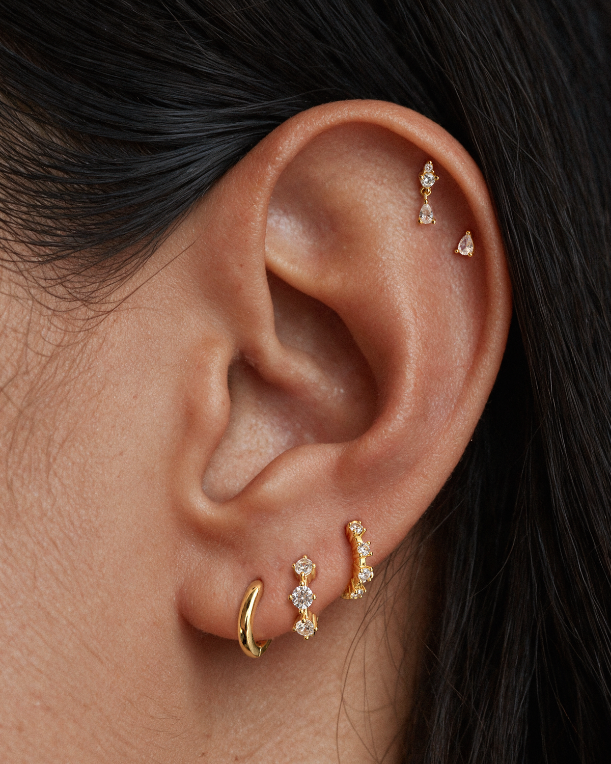 Close-up of a person's ear showcasing multiple 18k gold vermeil ear piercings, including a hoop, a curved huggie with clear stones, a marquise stud and one small dangling piercing stud in the upper helix.