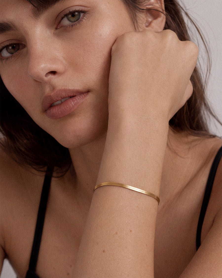 A close-up of a woman wearing a thin, polished 18k gold snake chain bracelet on her wrist, resting her head on her hand against a neutral light background.