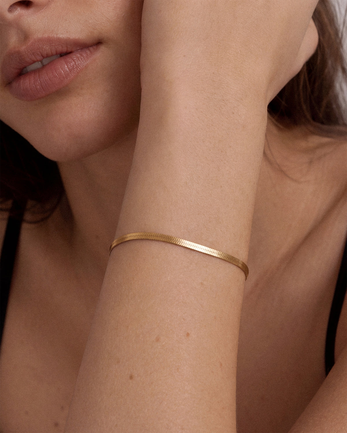 A close-up of a woman's wrist wearing a thin, polished 18k gold snake chain bracelet.