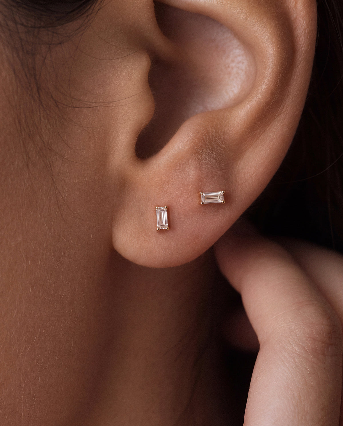 Close-up of a person's ear wearing two small rectangular 18k gold stud earrings in a warm metallic setting, with clear baguette stones.