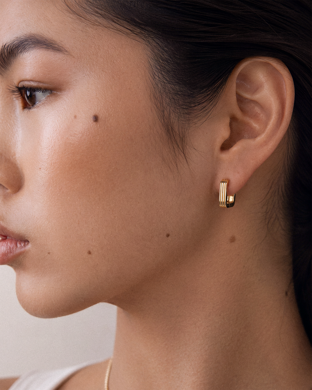 A close-up side profile of a woman wearing a small 18k gold hoop earring with vertical ridges.