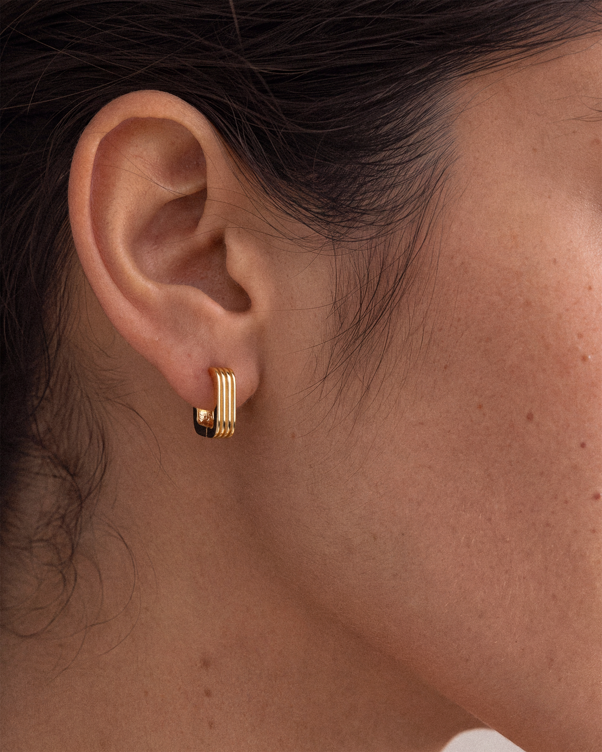 A close-up of a model's ear wearing a small 18k gold hoop earring with three parallel vertical bars.