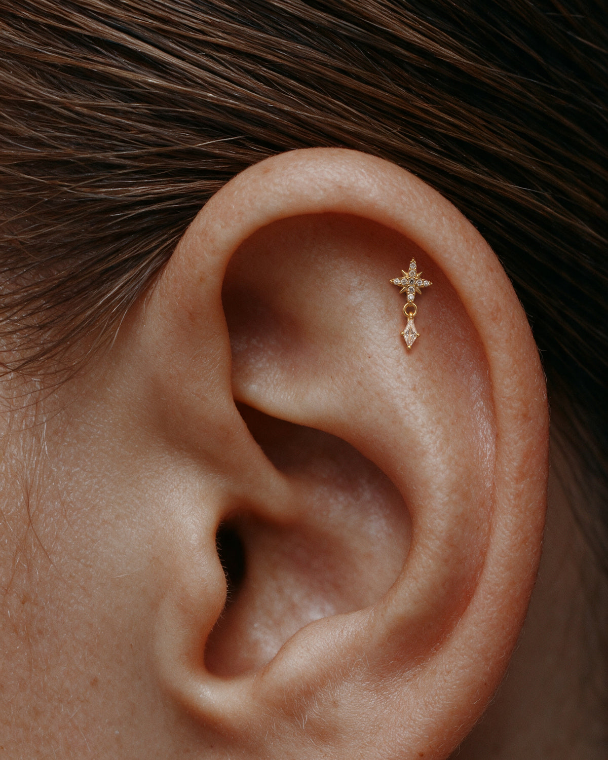 A close-up of a person's ear wearing a small 18k gold plated flatback ear stud with a starburst pave stud and a dangling diamond stone.