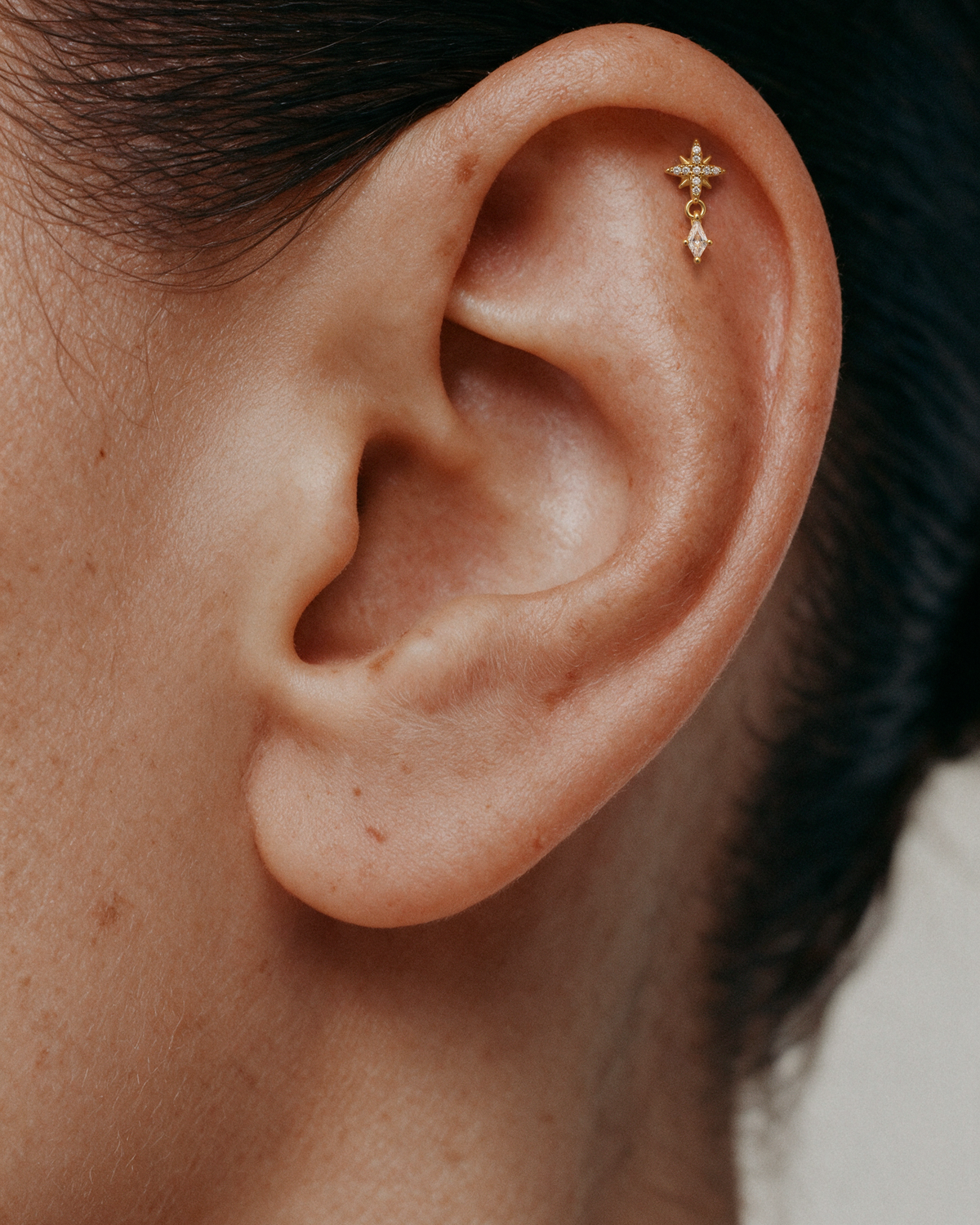 A close-up of a person's ear wearing a small 18k gold plated flatback ear stud with a starburst pave stud and a dangling diamond stone.