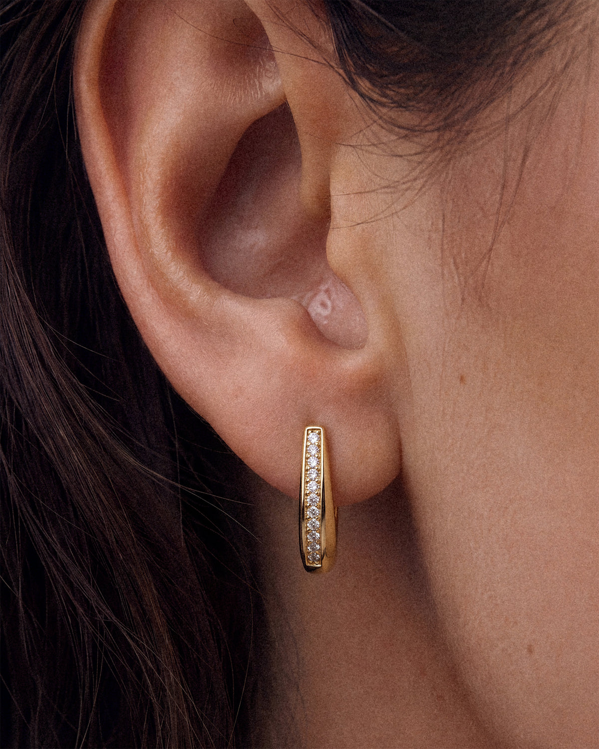 A close-up of a model's ear wearing an 18k gold hoop earring with a vertical channel of clear pavé stones and a tapered design.