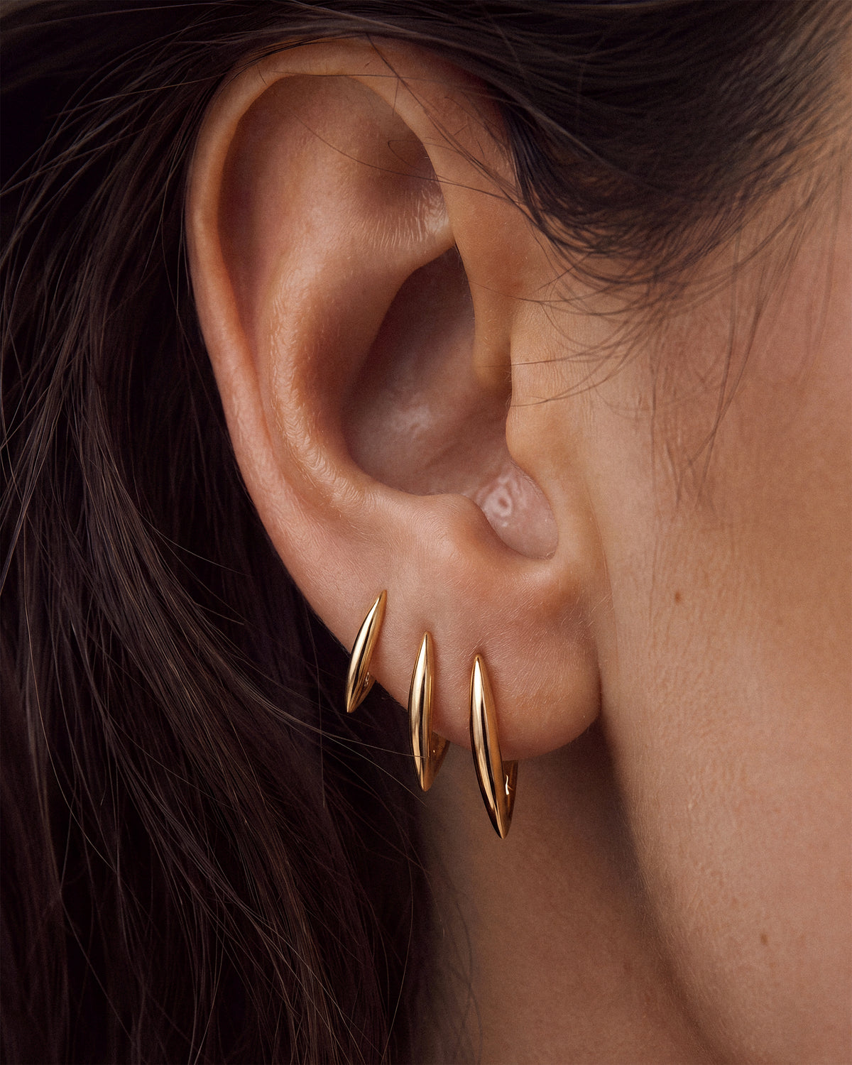 A close-up of a person's ear wearing three thin tapered 18k gold hoop earrings clustered on the lobe. 
