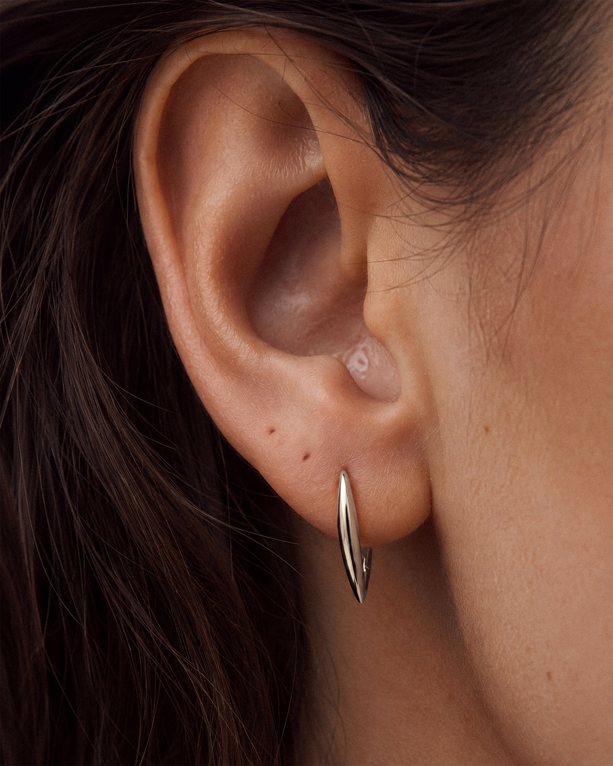 A close-up of a person's ear wearing a 925 sterling silver hoop earring with a polished triangular tapered shape/drop.