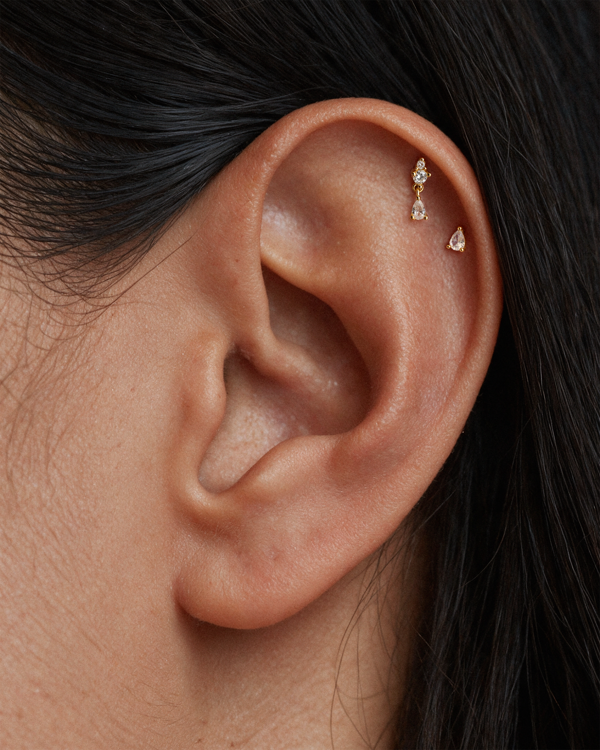 Close-up of a model's ear wearing two small 18k gold plated  ear stud, flatback earrings with clear gemstones on her upper helix piercings.