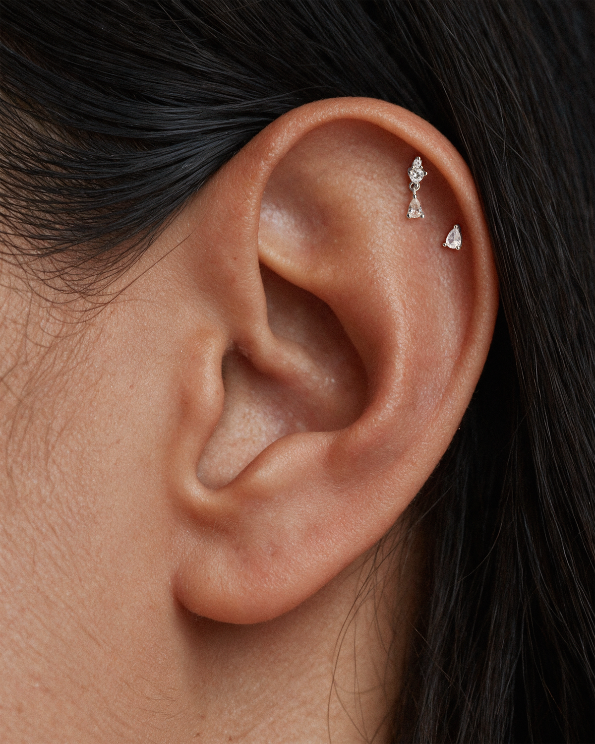 Close-up of a model's ear wearing two tiny 925 sterling silver ear stud, flatback earrings with clear gemstones on her upper helix piercings.