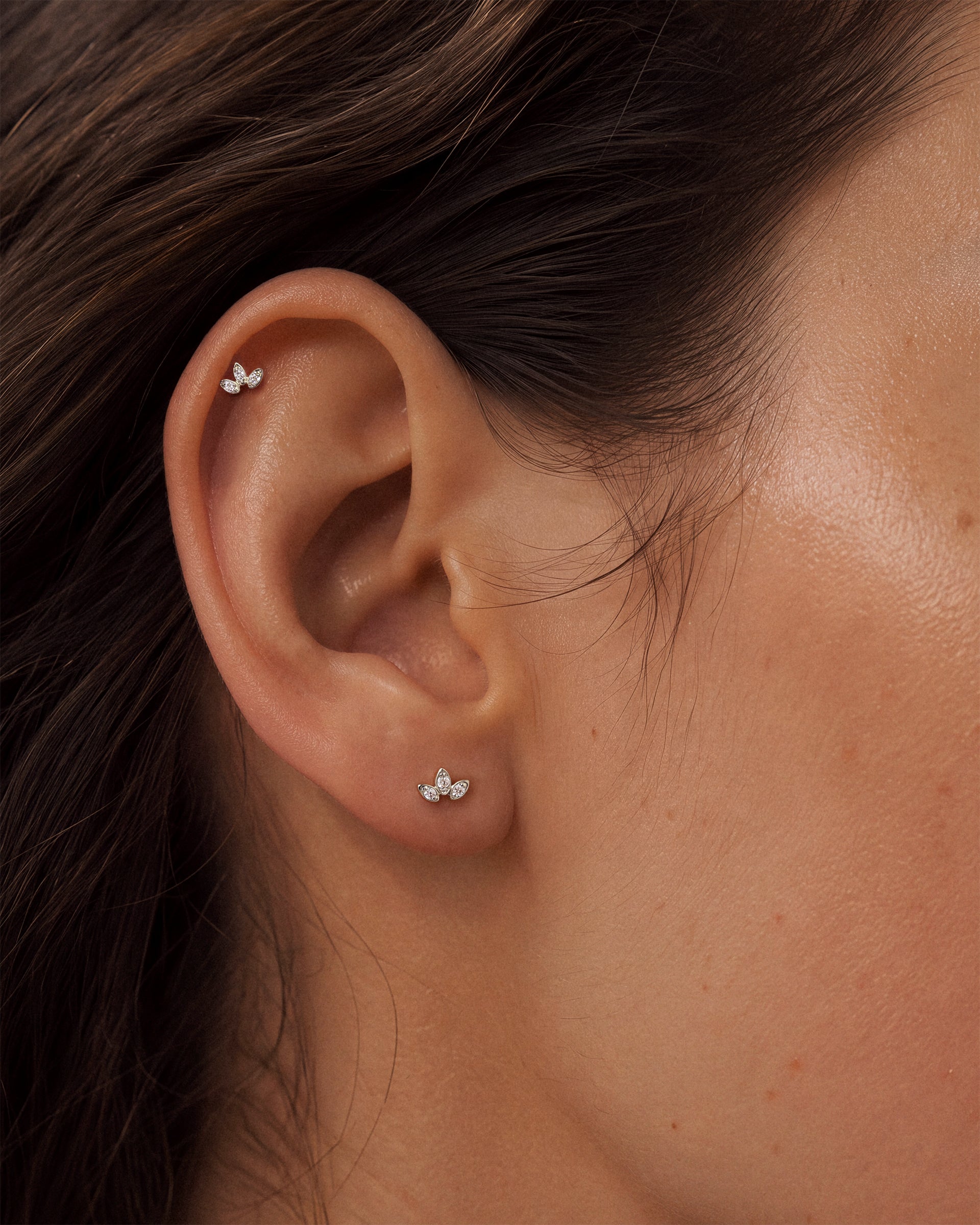 Close-up of a person's ear wearing two small 925 sterling silver stud earrings, one higher on the ear cartilage and one on the lobe, with dark brown hair and warm skin tones.