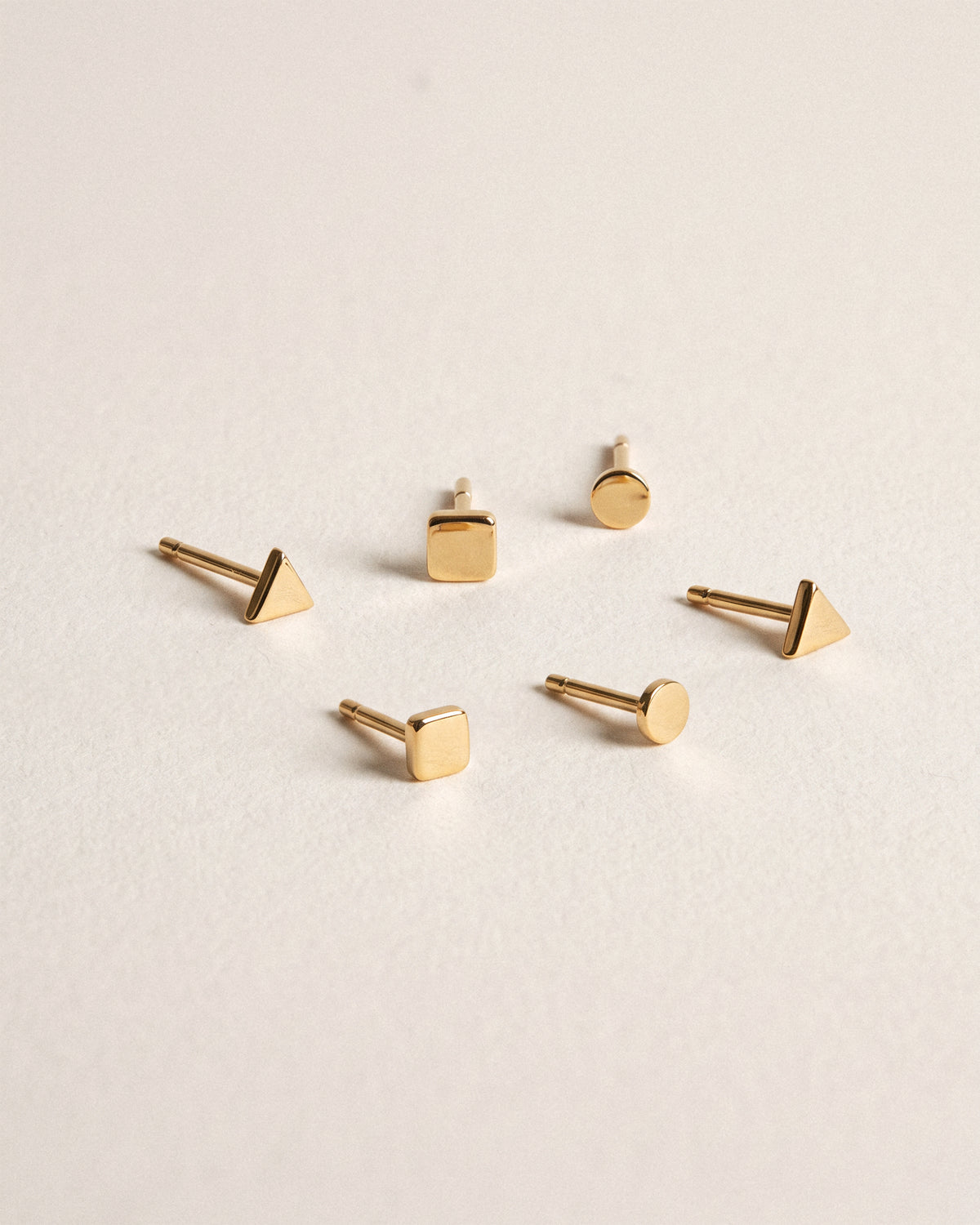 Six small 18k gold geometric stud earrings arranged on a light beige textured background, including triangle, square, and circle shapes. 