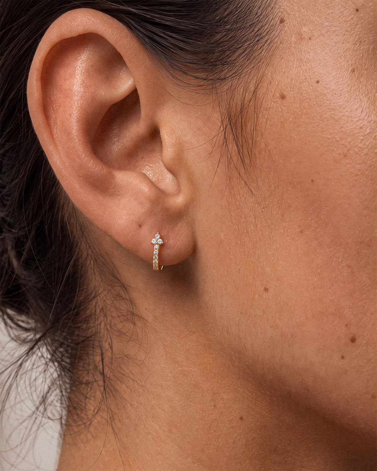 A close-up of a person's ear wearing a delicate 18k gold huggie hoop earring with a small cross at the top and cz gemstones.