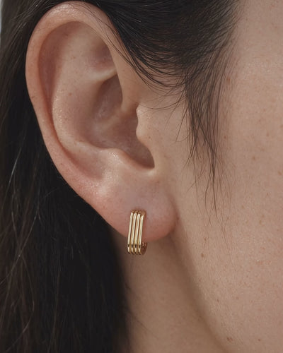 A close-up of a model's ear wearing a small 18k gold hoop earring with three parallel vertical lines/ridges.